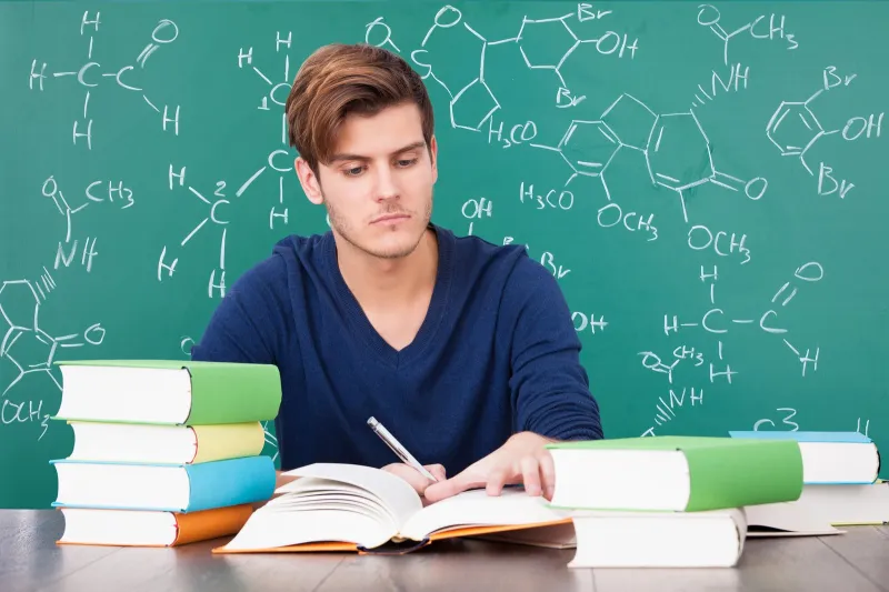 Maturzysta uczy się z podręcznika przed tablicą zapisaną wzorami strukturalnymi związków chemicznych